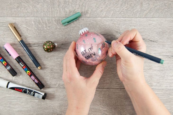 Eine Frau belettert eine rosa Christbaumkugel mit dem Spruch "Let ist snow" und Tannennadeln. Auf dem Tisch leigen diverse Marker der Marke Posca, Faber-Castell und Uniball.