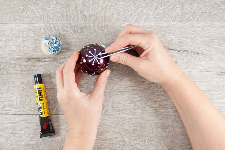 Eine Frau klebt auf eine beletterte Weihnachtskugel Glitzersteine mit einer Pnzette auf. Neben ihr auf dem Tisch liegt eine Tube UHU Kleber.