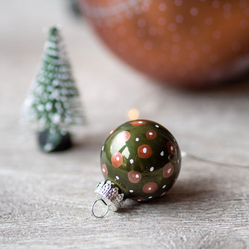 Nahaufnahme einer kleinen grünen Weihnachtkugel, die mit braunen und weißen Punkten belettert wurde. Im Hintergrund ist als Deko ein kleiner Tannenbaum zu sehen.