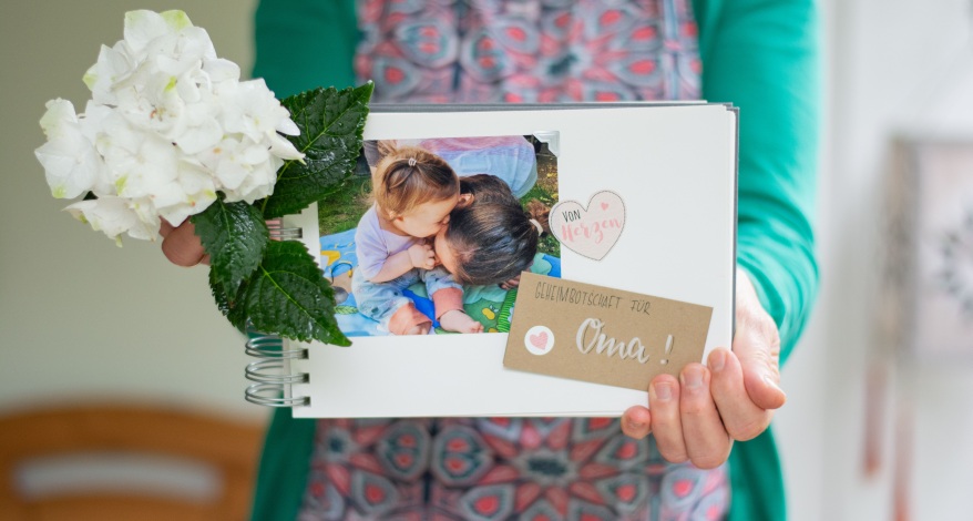 Eine Frau hält eine schön gestaltetes Fotobuch, in dem ein Kleinkind zu sehen ist, in die Kamera. In einer Hand hält sie noch einen Blumenstrauß.