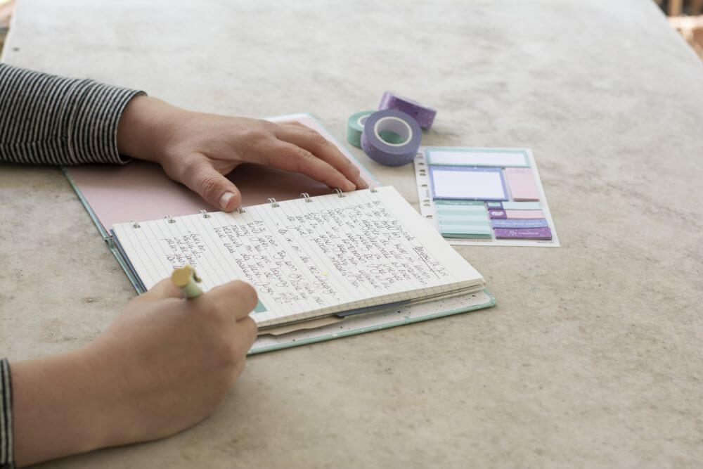 Ein Notizbuch von Filofax liegt aufgeklappt auf einem Schreibtisch, jemand schreibt hinein. Drumherum liegt ein Stickerbogen und Washitape.