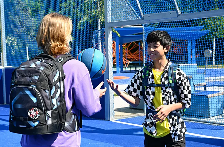 Zwei Jugendliche stehen mit ihren Schulrucksäcken von coocazoo auf einem Basketballplatz und werfen sich Bälle zu.