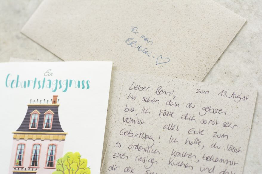 Auf einem Tisch liegt eine Geburtstagskarte mit Haus-Motiv und der Au7fschrift "Geburtstagsgruss". Daneben liegt eine aufgeklappte und handschriftlich geschriebene Karte sowie ein passender Umschlag.