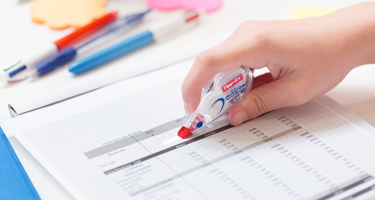 Hand korrigiert mit Tipp-Ex Mini Pocket Mouse eine gedruckte Tabelle auf einem Schreibtisch mit Stiften und Notizzetteln. 
