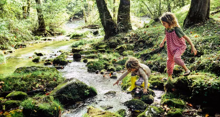 Zwei Kinder spielen an einem flachen Bach in einem Wald. Eines der Kinder kniet am Ufer und berührt das Wasser, während das andere danebensteht und neugierig zusieht. Beide tragen bunte, bequeme Kleidung und Gummistiefel. Um sie herum ist die Natur lebendig, mit Moos bedeckten Steinen, üppigem Grün und hohen Bäumen.