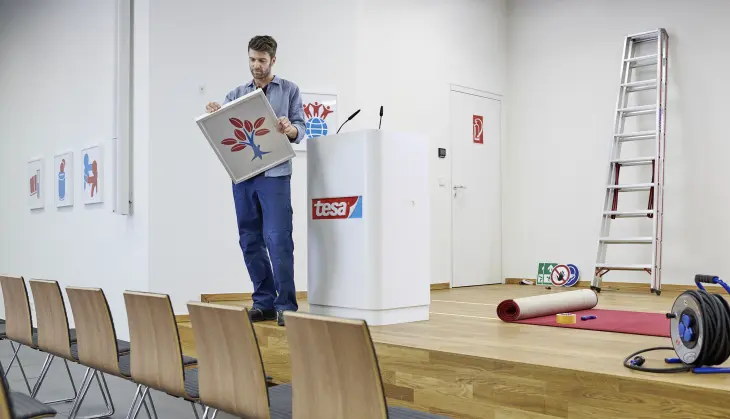 Ein junger Mann steht auf einer Bühne neben einem Podium mit tesa Logo und hält ein Bild in der Hand. Auf der Bühne sind unter anderem eine Leiter, ein aufgerollter Teppich und Reinigungsgeräte zu sehen. Vor der Bühne sind Holzstühle aufgestellt.