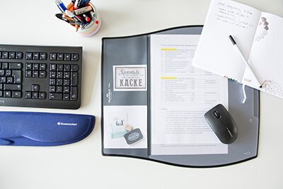 Aus der Vogelperspektive blickt man auf einen Arbeitsplatz. Man sieht eine Schreibtischunterlage mit Bildern und Notizen, eine Maus, ein Notizbuch, eine Tastatur, einen Stifteköcher und eine Handgelenkauflage.