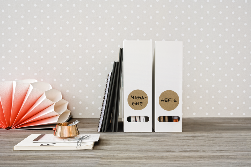 Auf einem Holztisch stehen 2 weiße Stehsammler, welche mit braunen runden Etiketten der Marke Avery Zweckform beklebt und mit der Aufschrift "Magazine" und "Hefte" beschriftet sind. Daneben liegen ein aufgeschlagenes Buch, ein Notizblock und ein Behälter mit Büroklammern.