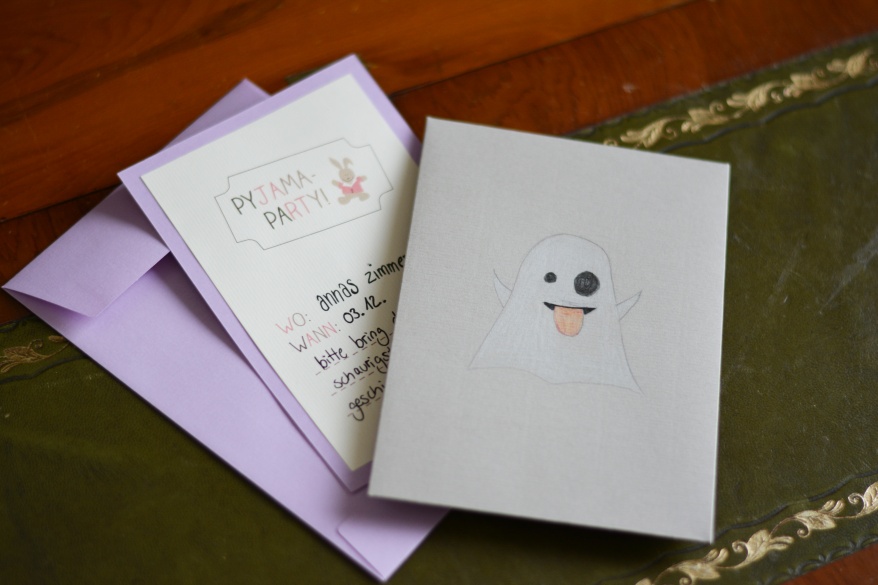 Eine selbstgestaltete Karte in lila mit einem gestaltetem Papier, auf dem eine Einladung zur Pyjamaparty steht und auf einem grauen Umschlag ist ein Gespenst gemalt.