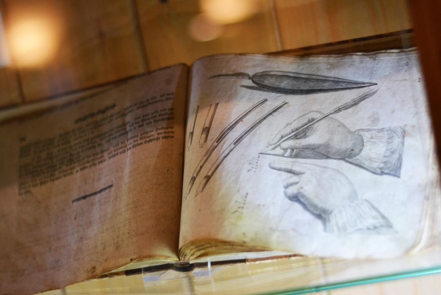 Ein altes Buch, aufgeschlagen auf einer Seite mit einer historischen Zeichnung zur Schreibfeder-Haltung.