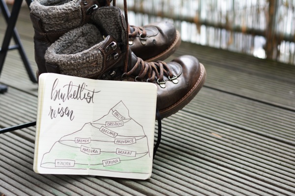 Wanderstiefel mit Notizbuch, das eine Reise-Bucketlist in Bergform zeigt.