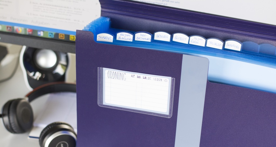 Registermappe mit Reitern vor Monitor, Aufschrift: „Ordnung ist das halbe Leben“.