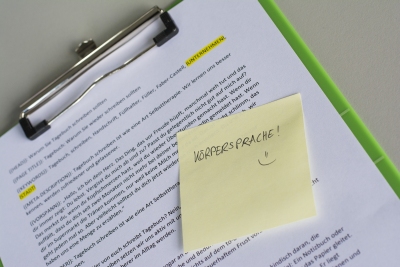 Klemmbrett mit Text und gelbem Post-it mit Notiz „Körpersprache! :)“