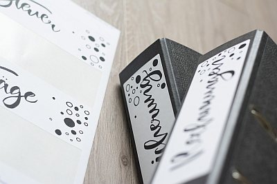 Auf einem Holztisch liegen zwei schwarze Ordneretiketten, die mit Etiketten der Marke Avery Zweckform beklebt wurden. Links davon liegt ein bedruckter Bogen mit Ordnerrückenetiketten, von dem zwei Etiketten entfernt wurden.