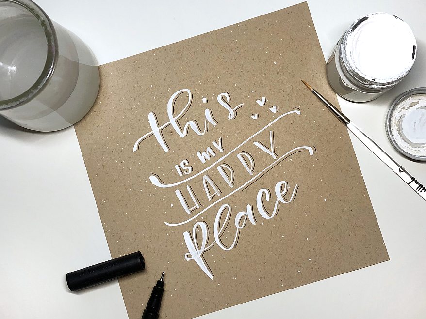 Auf einer braunen quadratischen Karte ist der Spruch "This is my happy place" in weißer Farbe gelettert. Daneben steht ein Glas mit Wasser und ein Glas mit weißer Aquarellfarbe. 