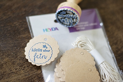 Auf einem dunklen Holztisch liegt eine Packung mit Geschenkanhängern von Heyda. Darauf liegt ein benutzter Stempel und ein einzelner Geschenkanhänger aus Kraftpapier. Der Anhänger wurde mit einem blauen Stempel bestempelt. Drauf ist zu lesen "klein aber fein".