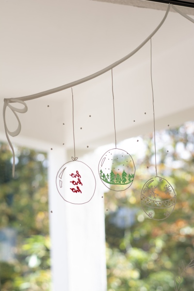 3 Weihnachtskugeln mit unterschiedlichen Motiven sind mit weißem Kreidemarker auf ein Fenster gemalt.