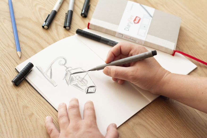 Zwei Händen skizzieren eine Straßenlaterne mit Stiften in Grautönen in ein Skizzenbuch von Hahnemühle.