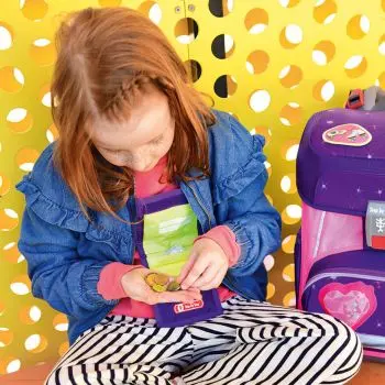 Ein rothaariges Mädchen sitzt vor einem gelben Hintergrund auf dem Boden. Neben ihr steht ein Schulranzen der Firma Step-by-Step. Sie hat einen Brustbeutel um den Hals hängen und zählt mit den Händen Kleingeld.