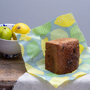 Auf einem kleinen Holztisch, der zur Hälfte mit einer beigen Leinentischdecke bedeckt ist, steht eine Keramikschüssel mit Zitrusfrüchten. Daneben liegt ein angeschnittenes Brot auf einem gemusterten Bienenwachstuch. Foto: Johanna Rundel