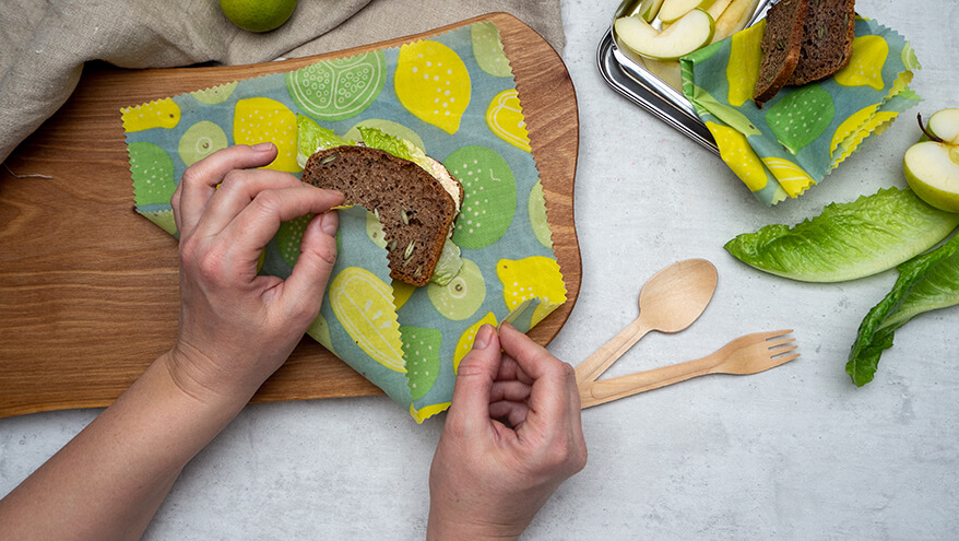 Jemand packt ein Pausenbrot in ein Bienenwachstuch ein, welches auf einem Holzbrett liegt. Daneben liegen auf dem Tisch Salatblätter, Apfelscheiben, Holzbesteck. Foto: Johanna Rundel