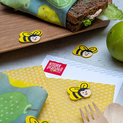 Auf einem hellgrauen Untergrund liegen Bienenwabenplatten von Knorr Prandell. Lustige Streudeko in Form von kleinen Holzbienen liegen darum verteilt. In der rechten unteren Ecke liegt lugt Holzbesteck in Bild. Am oberen Rand ist ein rustikales Holzbrett, auf dem ein, in ein Bienenwachstuch geschlagenes, Butterbrot liegt. Foto: Johanna Rundel