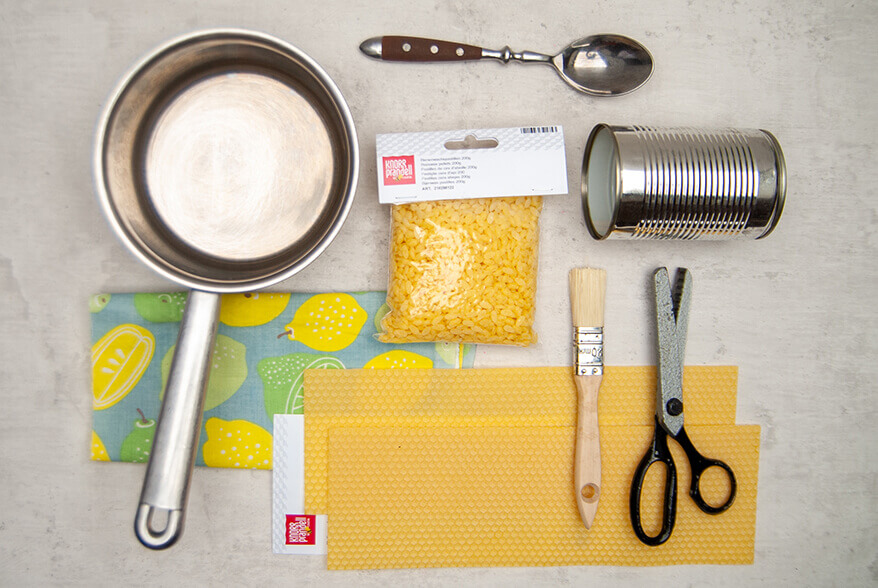 Auf einem hellgrauen Untergrund liegen die Materialien, die man zur Herstellung von DIY-Bienenwachtüchern benötigt: Topf, gemusterter Baumwollstoff, Bienenwachspastillen und -wabenplatten von Knorr Prandell, eine Blechdose, Pinsel, Zickzackschere und ein Esslöffel. Foto: Johanna Rundel