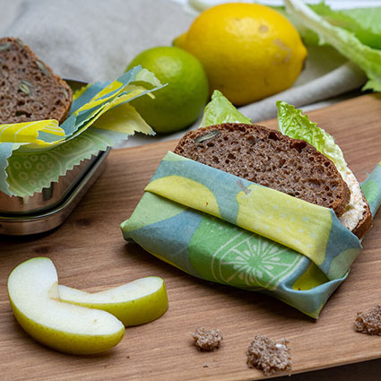 DIY_Bienenwachstuecher_Pausenbrot.jpg Ein Pausenbrot ist in ein gemustertes Bienenwachstuch eingeschlagen. Es liegt auf einem rustikalen Holzbrett. Drumherum sieht man Apfelstücke, Brotkrümel, eine Brotdose mit Brot und Zitrusfrüchte. Foto: Johanna Rundel