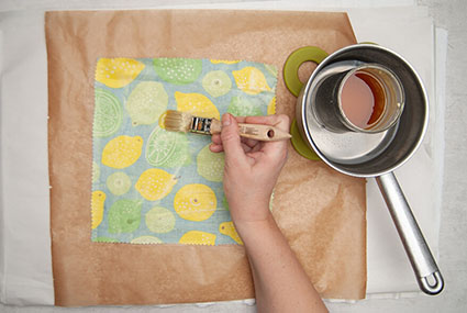 Ein quadratisches Stück gemusterter Stoff liegt auf einem Backpapier und wird mit einem Borstenpinsel mit flüssigem Wachs von Knorr Prandell aus einem Topf bestrichen. Foto: Johanna Rundel