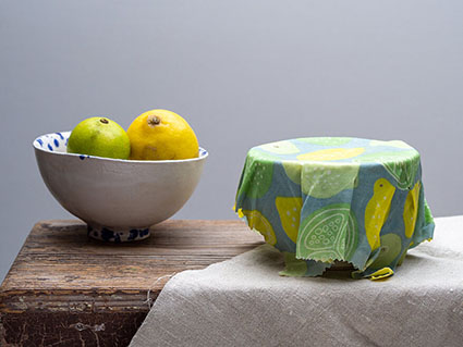 Auf einem kleinen Holztisch, der zur Hälfte mit einer beigen Leinentischdecke bedeckt ist, stehen zwei Keramikschüsseln. In der linken Schüssel liegen Zitrusfrüchte, die rechte ist mit einem gemusterten Bienenwachstuch abgedeckt. Foto: Johanna Rundel