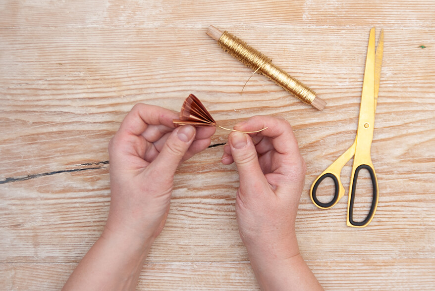 Auf einer hellen Holzoberfläche befestigt jemand goldenen Draht an einem zur Ziehharmonika gefaltetes Seidenpapier von Artoz. Daneben liegt eine Spule goldener Draht und eine goldene Schere. Foto: Johanna Rundel