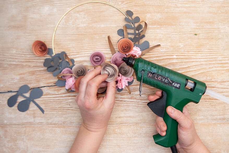 Auf einer hellen Holzoberfläche werden verschiedene Papierblumen und -blätter, die aus den Papieren von Artoz gefertigt wurden, mit Heißkleber auf einen goldenen Metallring geklebt. Foto: Johanna Rundel