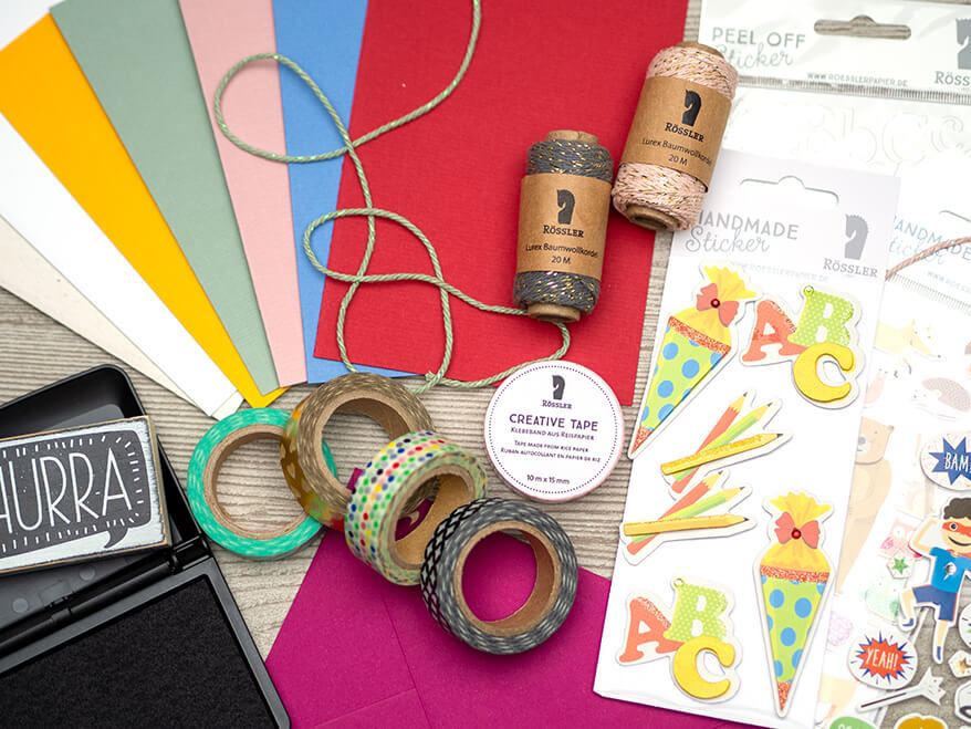 Auf einem hellgrauen Holzuntergrund liegen verschiedene Materialien und Papeterie der Marke Rössler: Blanko-Klappkarten, Washitapes, Stempel und Stempelkissen, Aufkleber, Sticker und Bäckergarn.
