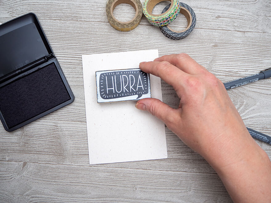 Auf einem hellgrauen Holzuntergrund liegt eine weiße Grußkarte der Marke Rössler. Darauf stempelt jemand mit schwarzer Farbe das Wort "Hurra".