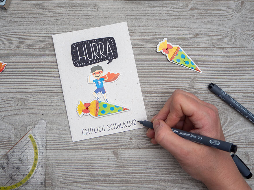 Auf einem hellgrauen Holzuntergrund liegt eine weiße Grußkarte der Marke Rössler. Jemand schreibt "Endlich Schulkind" unter zwei Sticker mit Schulmotiv und einem gestempelten "Hurra".