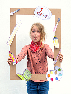 Ein Mädchen im Grundschulalter, mit blonden Haaren und roten Kleidern, hält einen Foto-Prop Bilderrahmen aus braunem Pappkarton vor sich in die Kamera. Auf dem Karton sind verschiedene Schulmaterialien aus Papier (Lineal, Stift, Buch etc.) zu sehen. Foto: Johanna Rundel
