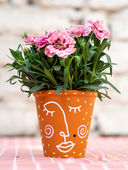 Auf einer rosa-weiß karierten Tischdecke steht ein Blumentopf aus Ton, der mit bunten Markern von Posca angemalt wurde. Auf dem Topf ist in Line-Art-Stil ein Frauengesicht gezeichnet. Im Topf befindet sich eine pinke Nelke. Im Hintergrund ist eine weiße Backsteinwand zu sehen. Foto: Johanna Rundel