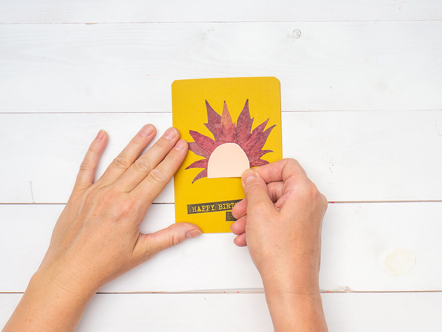 Jemand klebt einen Halbkreis aus rosa Tonkarton auf zwei getrocknete Herbstblätter, die auf eine gelbe Blanko Klappkarte von Artoz geklebt wurden. Auf die Karte wurde zuvor der Schriftzug „Happy Birthday“ gestempelt. Der Untergrund ist weiß. Foto: Johanna Rundel