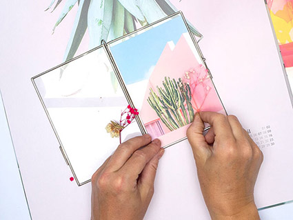 Jemand legt kleine, getrocknete Blumen auf eine Postkarte in einem Klapp-Bilderrahmen. Als Untergrund sind zwei Posterkalender von Athesia in Pastellfarben zu sehen. Foto: Johanna Rundel