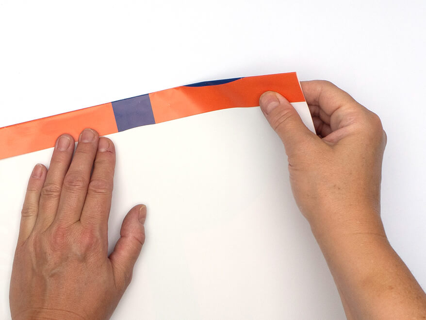 Tüte aus Papier falten ohne Vorlage: Jemand faltet ein rechteckiges, orangefarbenes Kalenderblatt von Athesia an der Längsseite einen Zentimeter um. Der Untergrund ist weiß. Foto: Johanna Rundel 