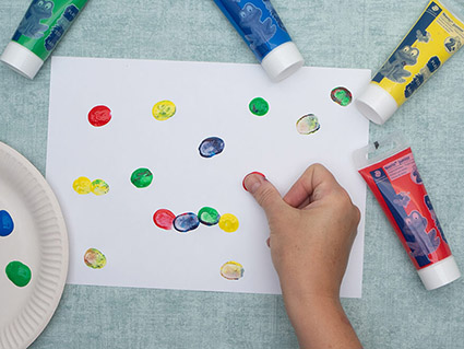 Jemand drückt mit Fingermalfarben von Staedtler bunte Fingerabdrücke auf ein weißes Blatt Papier. Der Untergrund ist türkis. Foto: Johanna Rundel"