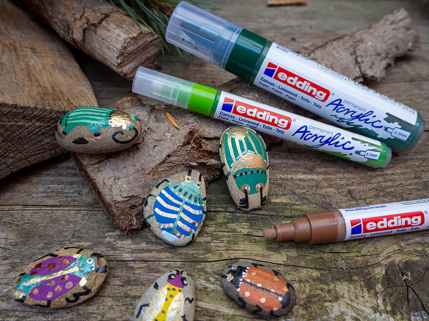 Auf einem rustikalen Holzuntergrund liegen mehrere Steine, die als Käfer bemalt wurden. Darüber sieht man die dafür verwendeten Acrylmarker der Marke edding. Foto: Johanna Rundel
