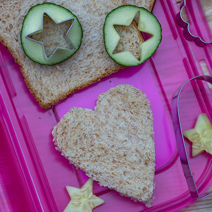Auf dem Deckel einer pinken Brotdose von School-Mood liegt ein in Herzform ausgestochenes Toastsandwich, sowie Gurkenscheiben aus denen mittig Sterne ausgestochen wurden. Die Plätzchenausstecher liegen daneben. Foto: Johanna Rundel