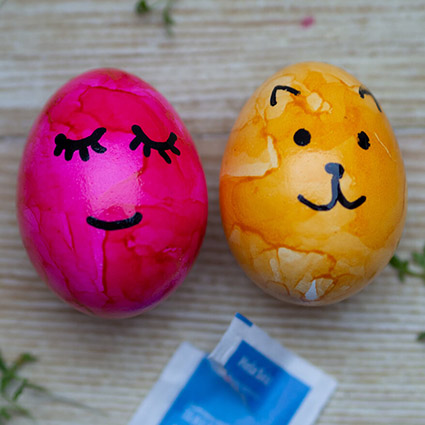 Auf einem Holzbrett liegen ein pink und ein gelb gefärbtes, gekochtes Ei, auf deren Schale mit schwarzem Stift ein Gesicht gemalt wurde. Untendrunter ist ein blaues Tütchen mit Salz zu sehen. Foto: Johanna Rundel