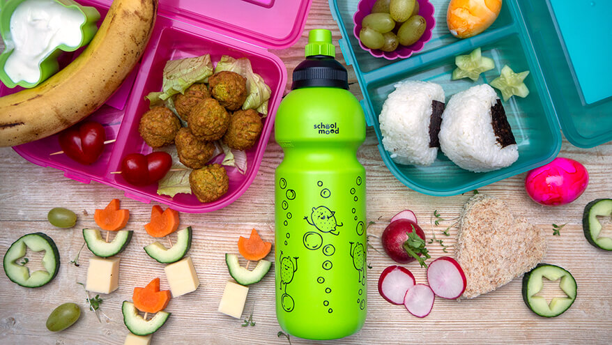 Auf einem Holzbrett liegen eine pinke und eine türkise Schulbrotdose sowie eine grüne Trinkflasche der Marke School-Mood. Darin und drumherum sind alle möglichen Speisen für gesunde Pausensnacks verteilt: Joghurt, Banane, Falafelbällchen, Käsespieße, Radieschen, Reispäckchen und Weintrauben. Foto: Johanna Rundel