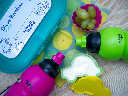 Auf einem Holzbrett liegen eine pinke und eine grüne Trinkflasche von School-Mood. Außerdem sind eine türkise Brotdose aus Plastik, eine kleine Plastikaufbewahrungsdose mit Deckel in Autoform, in der Joghurt ist sowie ein pinkes Silikon Muffinförmchen mit grünen Weintrauben zu sehen. Foto: Johanna Rundel