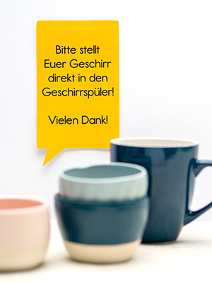 An einer weißen Wand hängt ein gelbes Hinweisschild von Avery-Zweckform in Form einer eckigen Sprechblase. Darauf steht: „Bitte stellt Euer Geschirr direkt in den Geschirrspüler. Vielen Dank!“. Davor sind – unscharf – verschiedene Tassen und Kaffeebecher zu sehen. Foto: Johanna Rundel