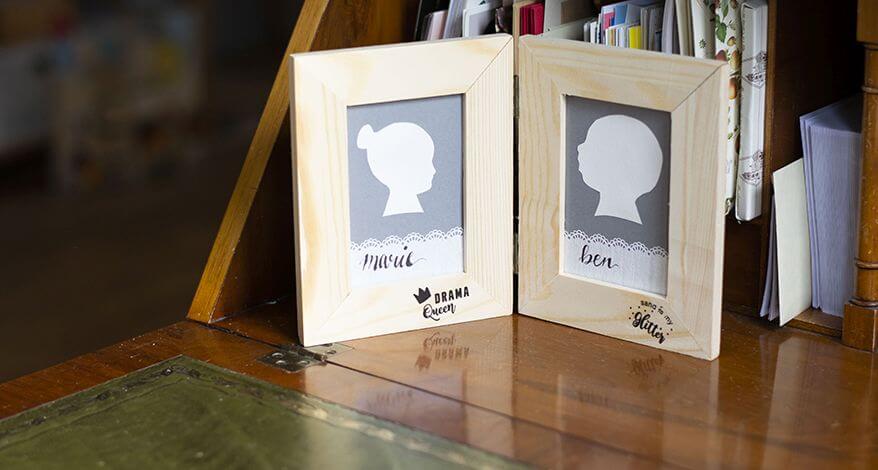 Ein Zweifachbilderrahmen mit zwei Do-it-yourself-Scherenschnitten von einem Jungen und einem Mädchen steht auf einem antiken Sekretär. Die cremeweißen Scherenschnitte sind auf einem grauen Hintergrund aufgeklebt. Darunter wurden auf einem Stück selbstklebenden Spitzentape im Handlettering-Stil die Namen Marie und Ben geschrieben. Auf den Bilderrahmen sind Rubbelsticker der Marke Knorr Prandell aufgebracht, auf ihnen steht „Drama Queen“ und „Sand is my glitter“.
