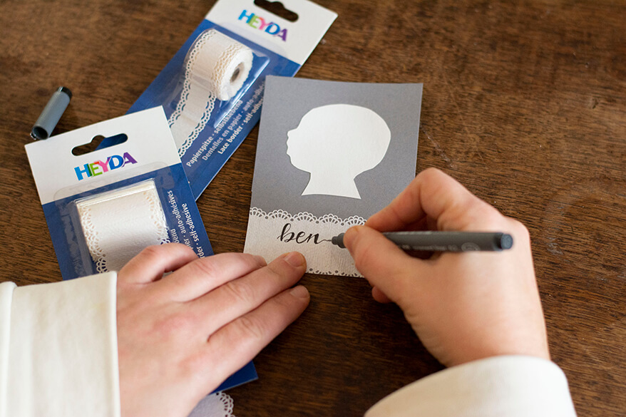 Eine Hand schreibt auf ein Stück selbstklebendes Spitzentape der Marke Heyda im Handlettering-Stil den Namen Ben. Es klebt auf einem Stück grauen Fotokarton, darüber ist ein cremeweißer Scherenschnitt eines Kinderkopfes zu sehen.