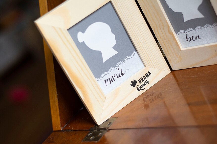 Man sieht einen Zweifachbilderrahmen mit Do-it-yourself-Scherenschnitten im Anschnitt auf einem antiken Sekretär stehen. Sichtbar ist der cremefarbene Kopf eines Mädchens im Profil. Der Scherenschnitt ist auf einem Stück grauem Fotokarton aufgeklebt, darunter steht auf einem Stück Spitzentape im Handlettering-Stil der Name „Marie“. Auf dem Bilderrahmen wurde ein Rubbelsticker der Marke Heyda aufgetragen, auf ihm steht „Drama Queen“.